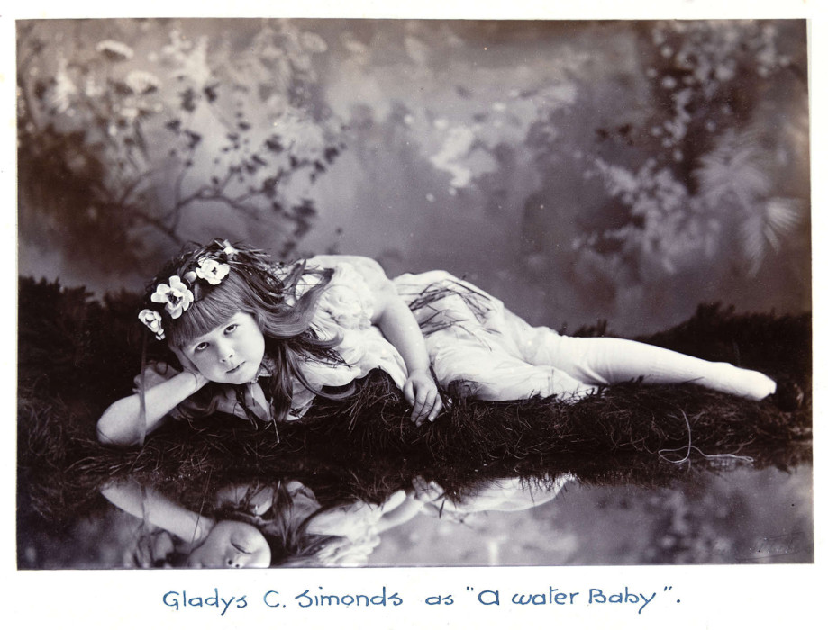 A girl lies down on water's edge looking to camera. Words 'Gladys C. Simonds as A Water Baby' written on bottom Ref. D/E1792/1